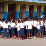 En avant plan de droite à gauche en chemise verte un enseignt en conversation avec des élèves habillés en bleu blanc devant le batiment d'une école à Kinshasa Radio Okapi Ph John Bompengo