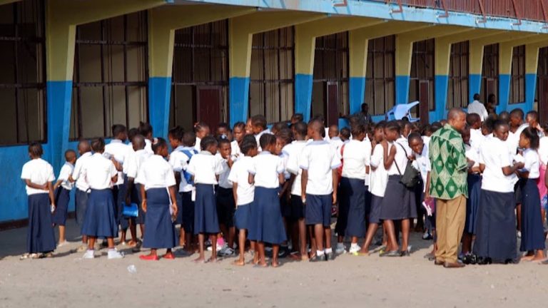 En avant plan de droite à gauche en chemise verte un enseignt en conversation avec des élèves habillés en bleu blanc devant le batiment d'une école à Kinshasa Radio Okapi Ph John Bompengo